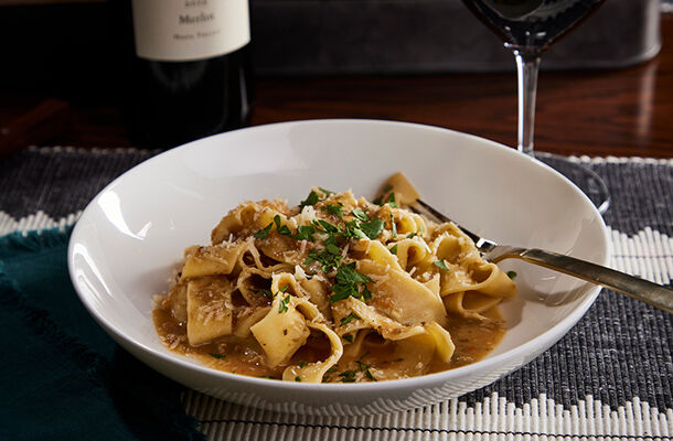 Pappardelle pasta with vegetable Bolognese sauce served in a white bowl, garnished with fresh herbs, with a glass of red wine.