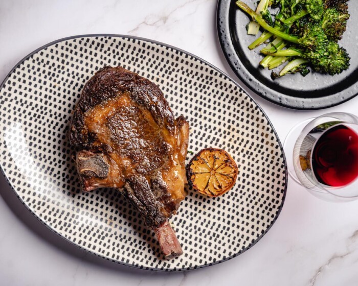 Seared bone-in ribeye steak with roasted broccolini and a glass of red wine on a white marble surface.