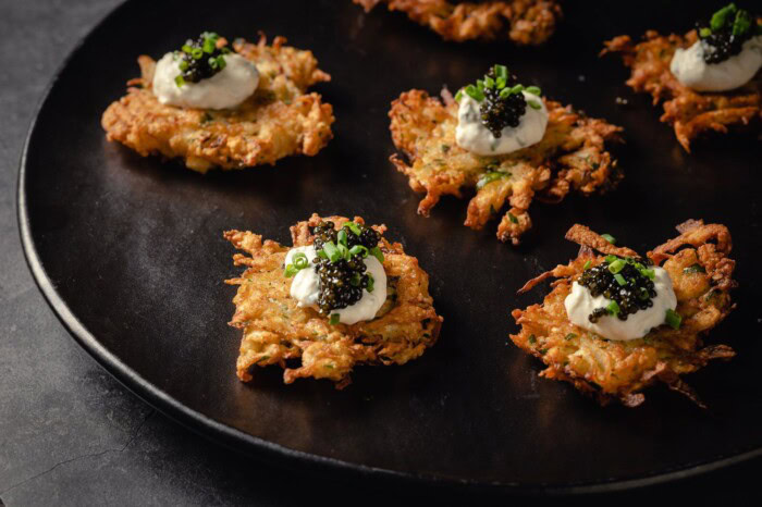 Crispy celery root latkes topped with horseradish sauce and caviar on a black plate, served as a gourmet appetizer.