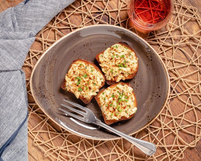 Creamy deviled crab toasts served on a rustic gray plate with a fork, garnished with herbs, and accompanied by a glass of ros.