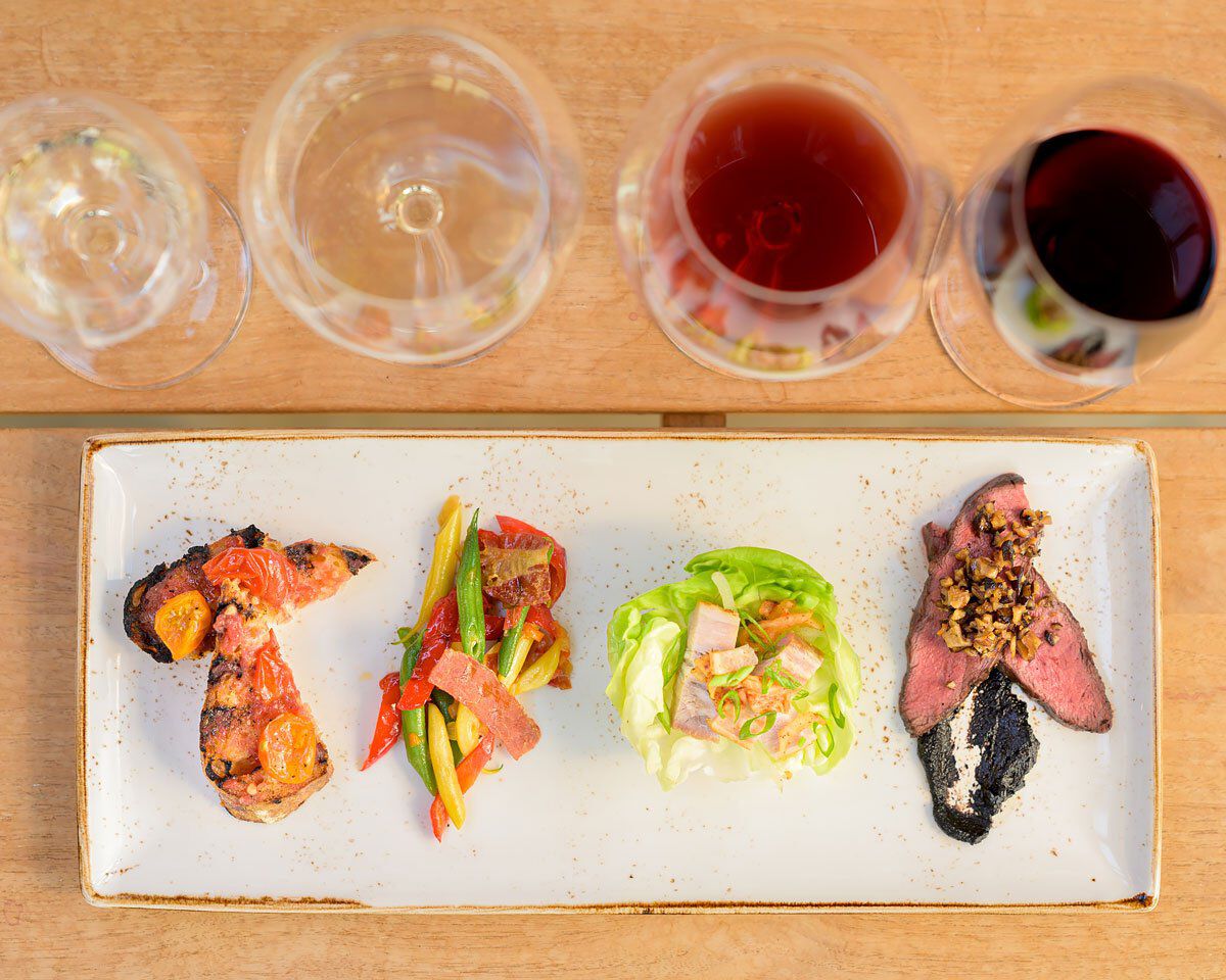 A detailed tasting plate with grilled meat, vegetable salad, lettuce wrap, and sliced beef, paired with three glasses of red.
