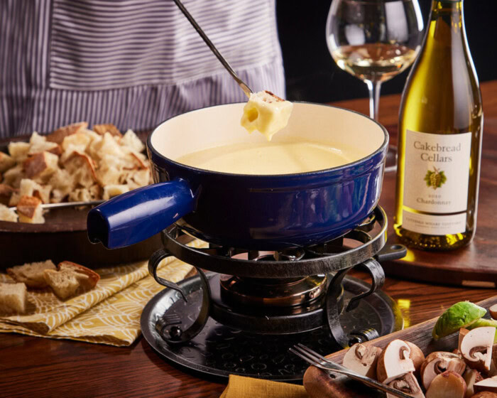 Creamy cheese fondue in a blue pot with a fondue fork, surrounded by bread, mushrooms, and a bottle of Cakebread Cellars wine.