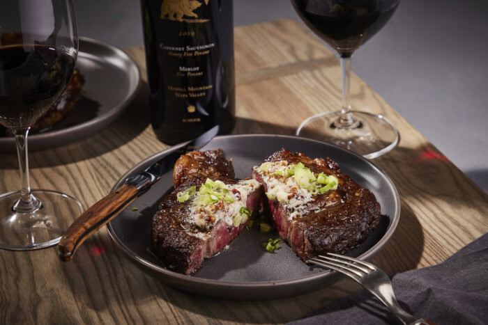 Juicy ribeye steak with pecan, bourbon, and blue cheese butter served on a black plate with wine glasses and a bottle in the.