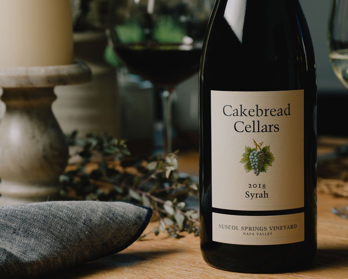 Cakebread Cellars Syrah wine bottle on a wooden table with a glass of red wine in the background.