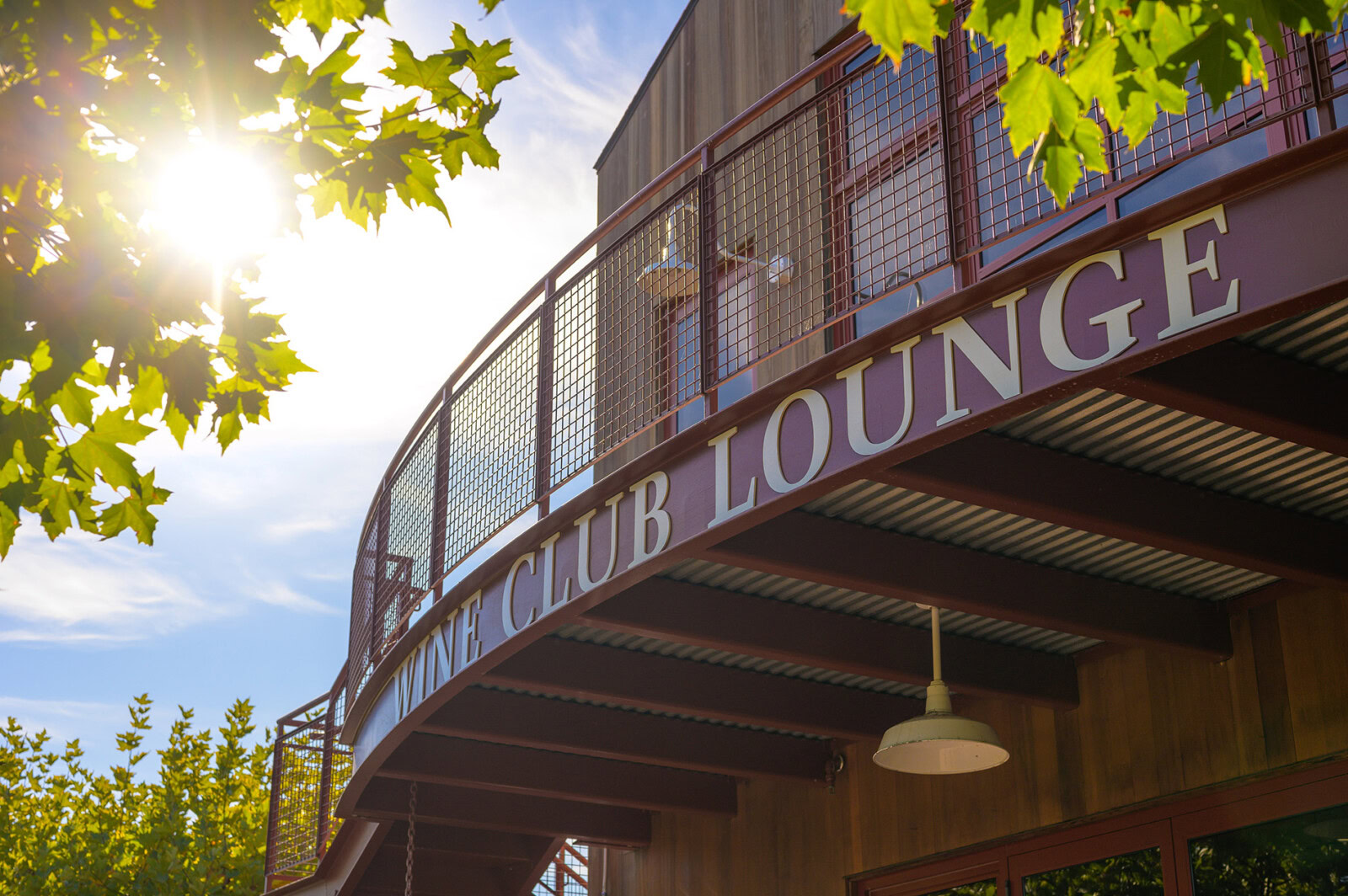 A lush outdoor view of Cakebread Cellars' wine club lounge with a curved balcony and hanging light, surrounded by green trees.