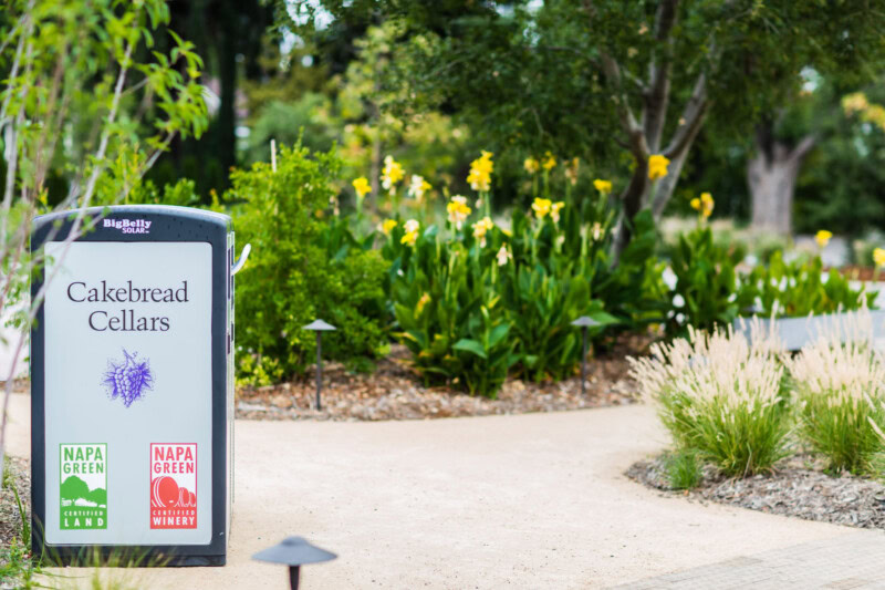 Cakebread Cellars signboard outdoors with lush greenery and yellow flowers in the background.