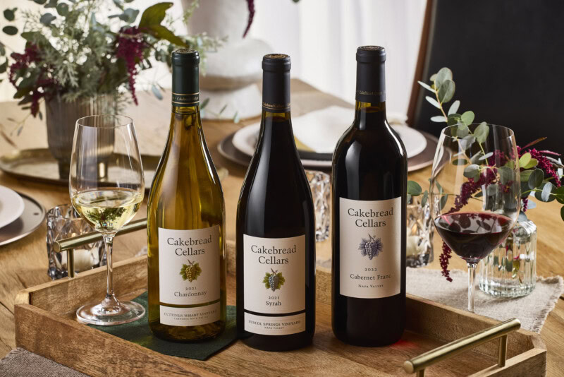 A lush display of three Cakebread Cellars wine bottles with glasses of white and red wine on a wooden tray, set on a table wi.