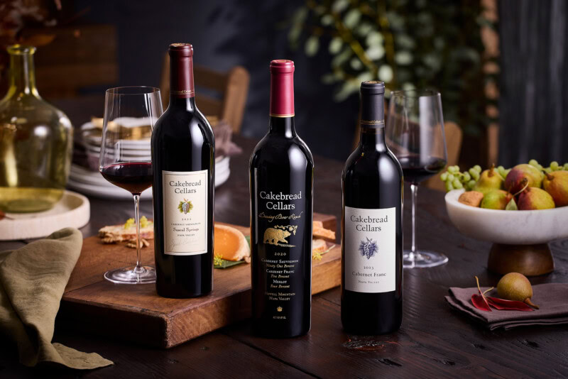 A lush display of three Cakebread Cellars wine bottles with glasses on a wooden table, surrounded by grapes and a cozy, elega.