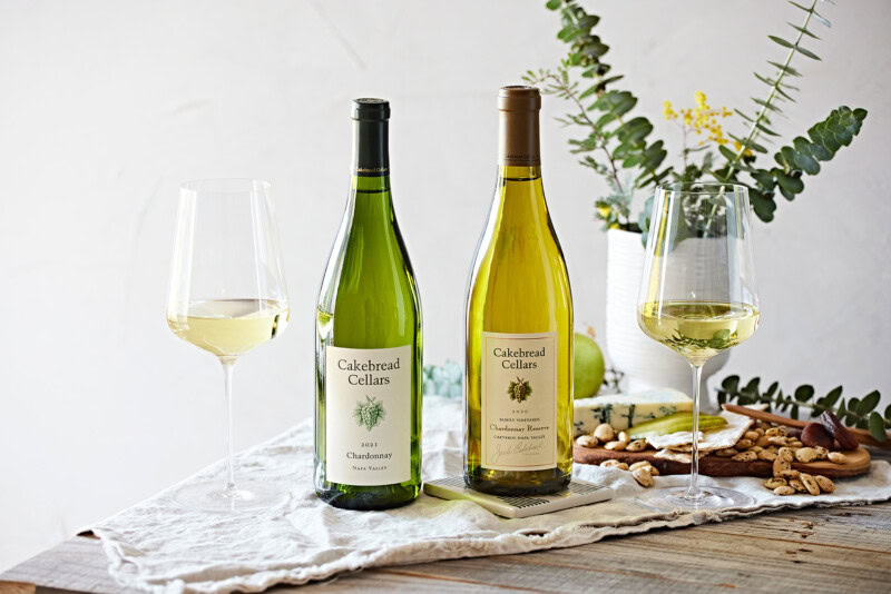 Two bottles of Cakebread Cellars Chardonnay with wine glasses on a rustic table, fresh greenery, and snacks in the background.