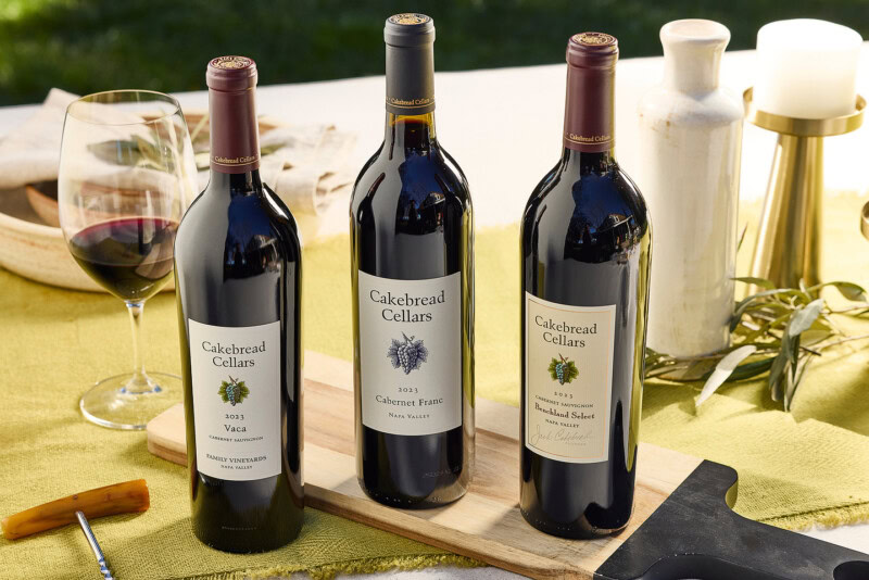 Three Cakebread Cellars wine bottles on a wooden board with a glass of red wine, candles, and greenery in the background.
