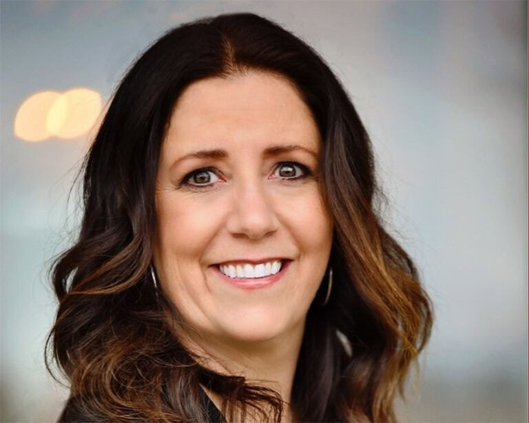 Gina Charbonneau with long, wavy brown hair smiling, standing outdoors with a blurred background.