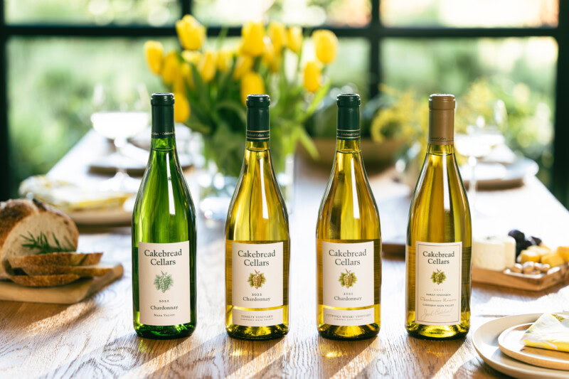 A lush display of four Cakebread Cellars Chardonnay wine bottles on a wooden table with a vase of yellow tulips in the backgr.