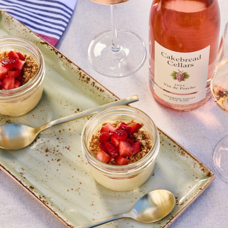 Creamy dessert in glass jars topped with fresh strawberries, served on a rustic ceramic tray with a bottle of Cakebread Cella.
