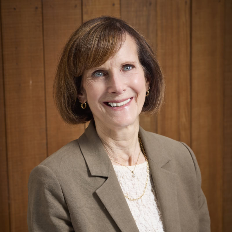 Professional woman smiling, wearing a beige blazer and white top, in front of a wooden background.