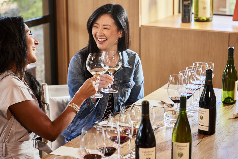 Two women enjoying wine tasting at Cakebread Cellars with wine bottles and glasses on the table.