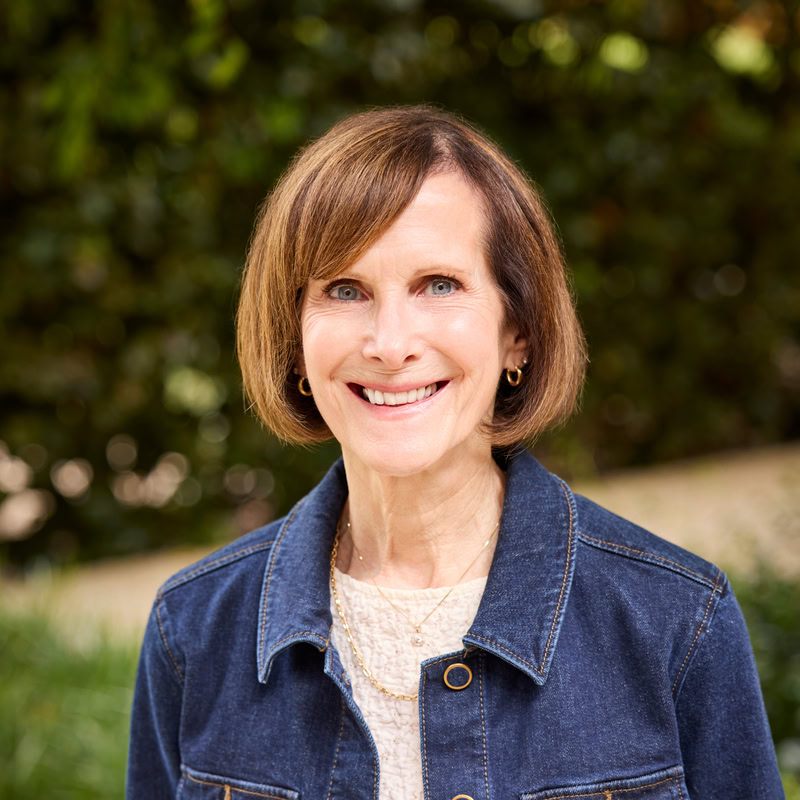 Smiling woman with short brown hair wearing a denim jacket outdoors.