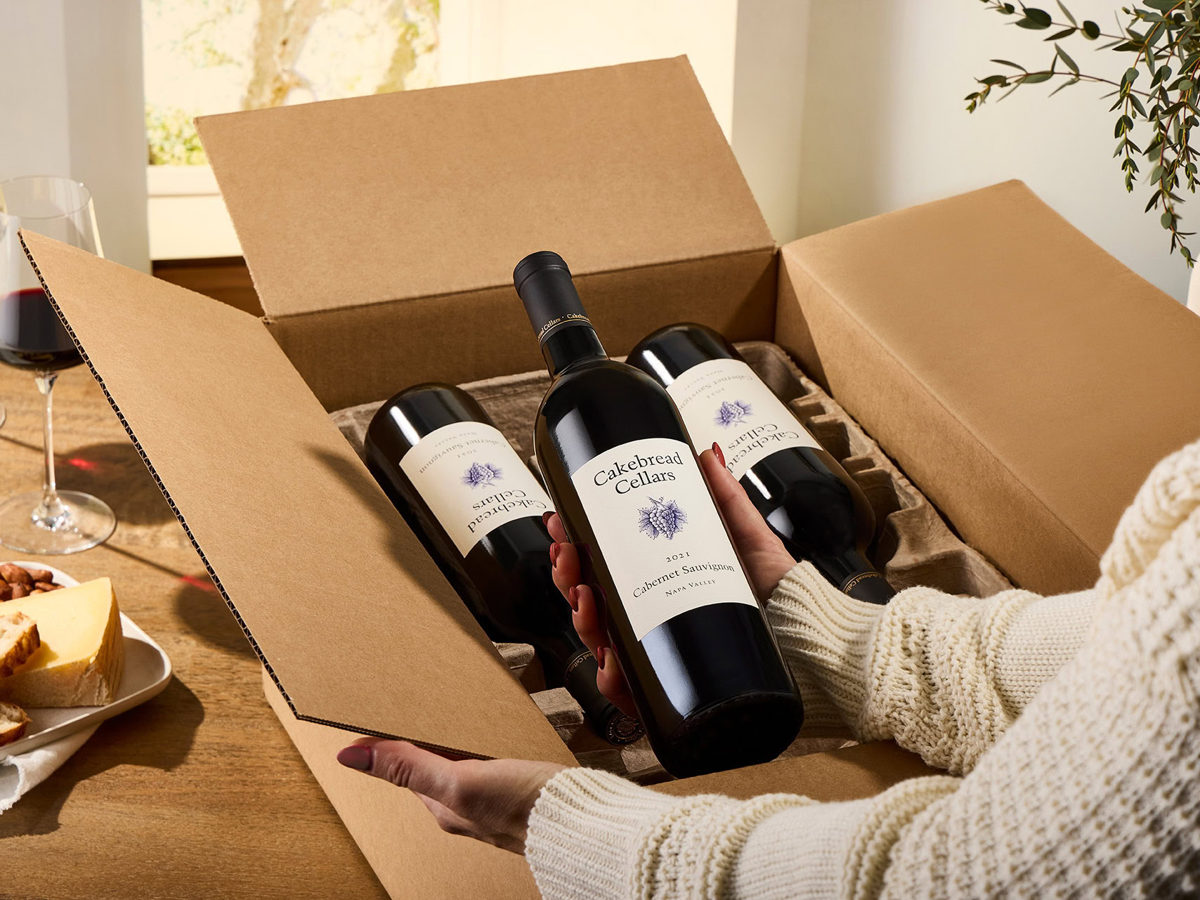 A woman holding three bottles of Cakebread Cellars wine in a cardboard box.