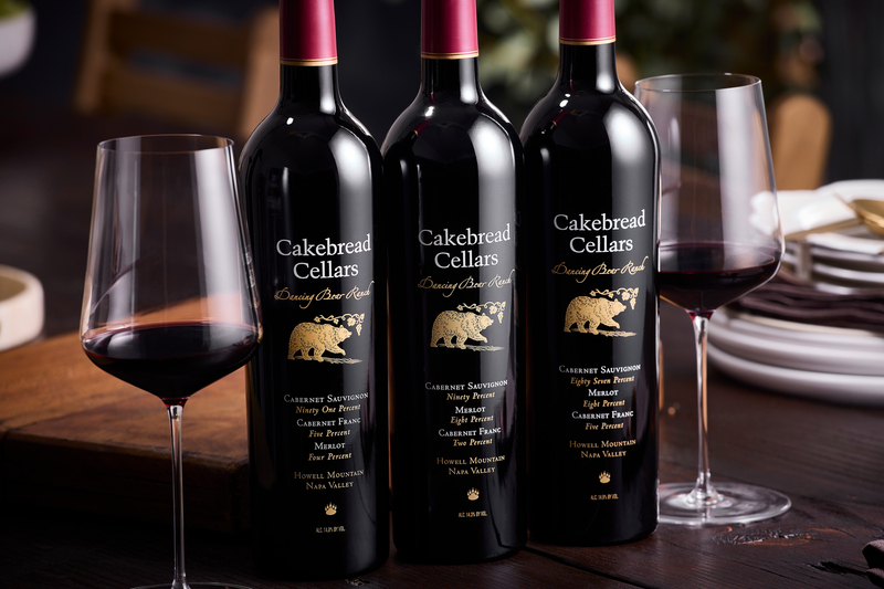 Three bottles of Cakebread Cellars wine with two glasses of red wine on a dining table.