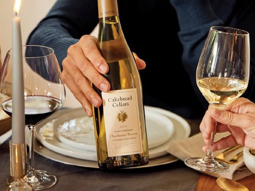 Person holding a bottle of Cakebread Cellars Chardonnay with wine glasses on a dinner table.